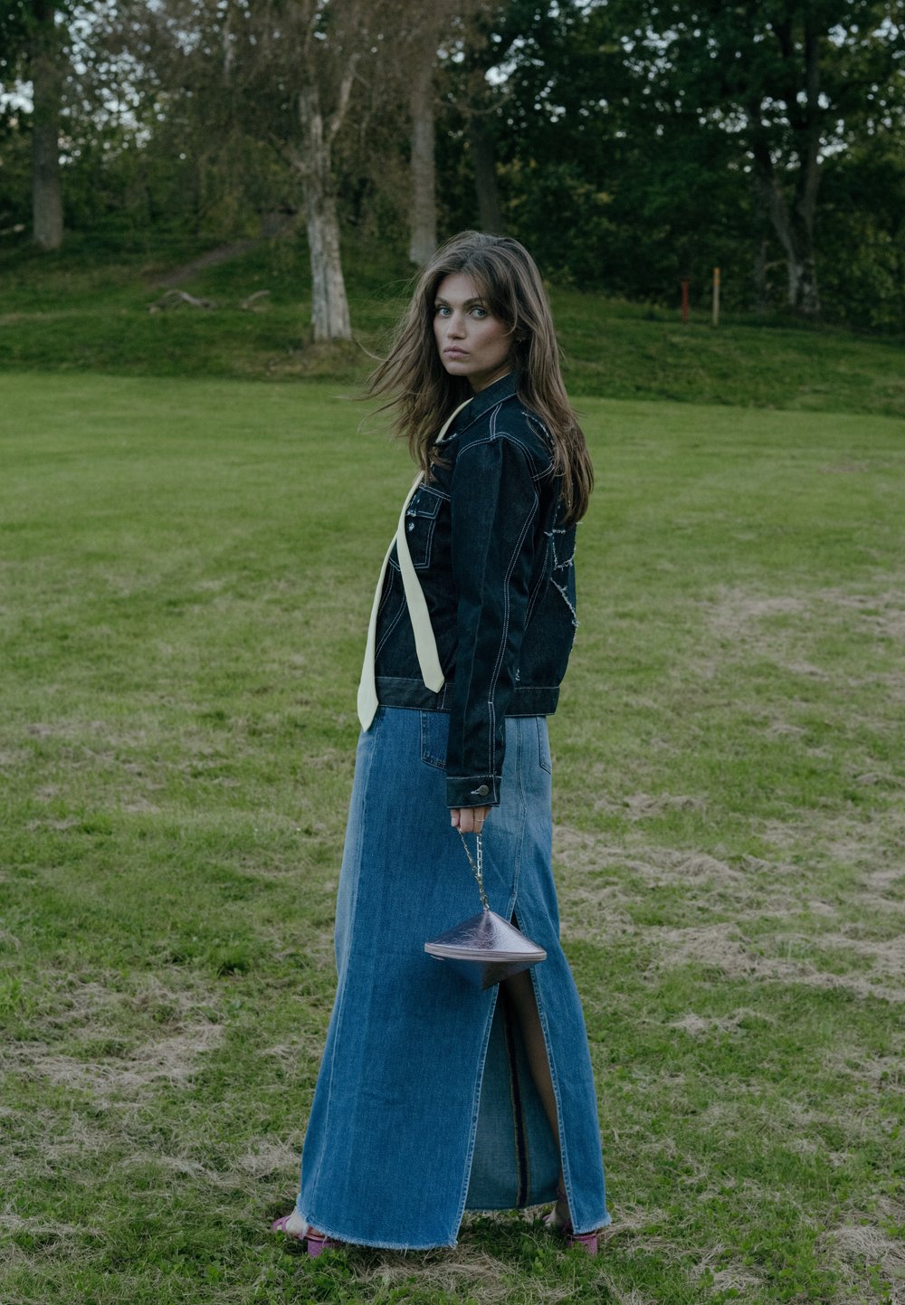 Dżinsowa kurtka z akcentami przyszycia, w zestawieniu z długą spódnicą dżinsową z rozcięciem. Dodatki obejmują małą, błyszczącą, fioletową torebkę.