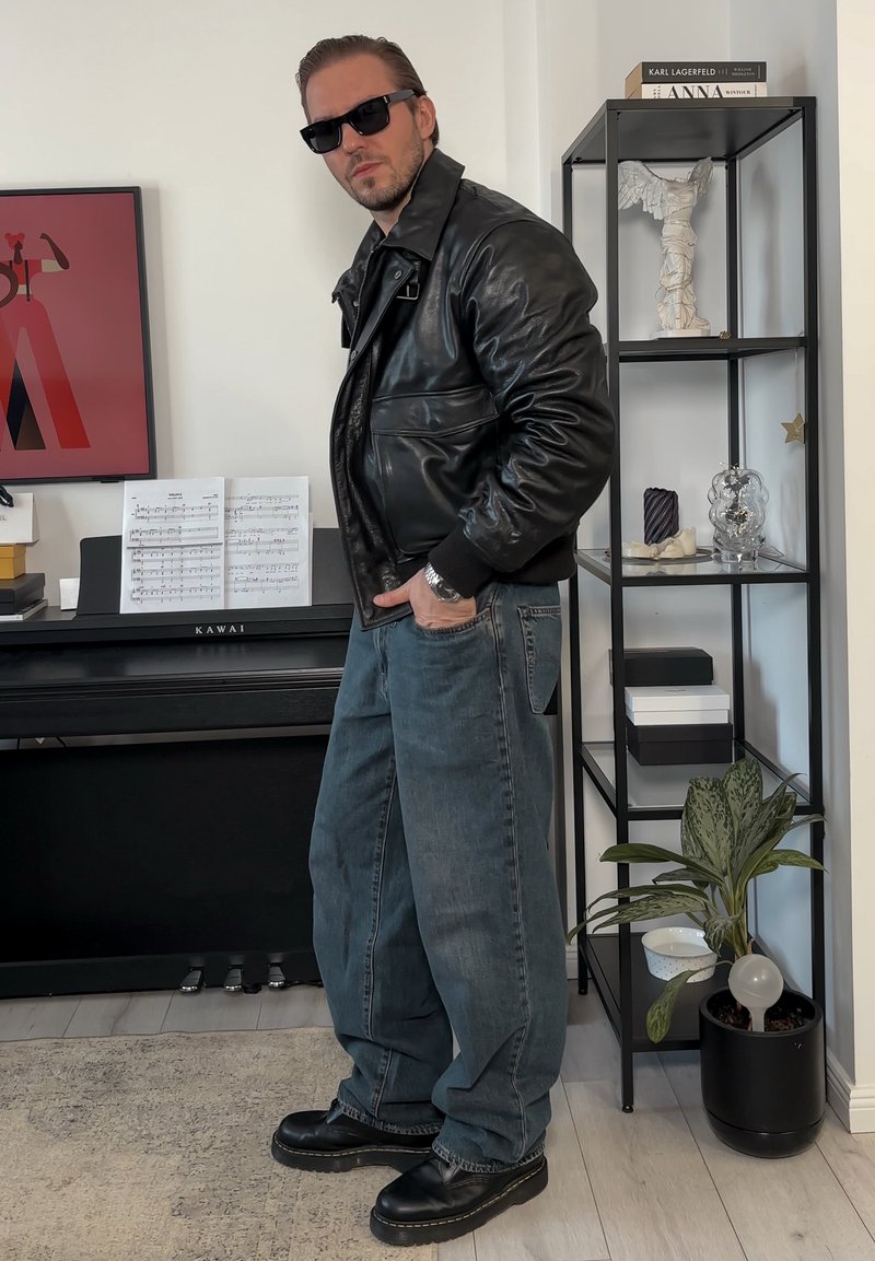 Veste aviateur en cuir noir, pantalon large en jean bleu, chaussures noires, et lunettes de soleil. Fond neutre avec un piano et une étagère décorative.