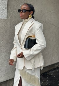 Γυναίκα με λευκό κοστούμι με φουντίτσες, μαύρα γυαλιά ηλίου, μεγάλα χρυσά σκουλαρίκια, κρατώντας μαύρη και χρυσή τσάντα, όρθια δίπλα σε υφή τοίχου.