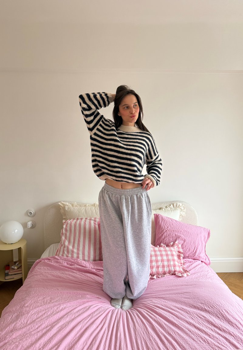 Femme en pull rayé et pantalon de survêtement gris debout sur un lit rose avec des oreillers rayés et unis dans une chambre minimaliste.