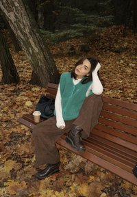 Zielona kamizelka polarowa na białej koszuli z długim rękawem, brązowe spodnie szerokie, czarne buty. Drewniana ławka z opadającymi liśćmi i filiżanką kawy.