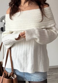 Femme portant un pull côtelé blanc à épaules dénudées, une jupe en jean claire, des collants noirs, tenant un sac à main en cuir marron à l'intérieur.