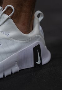 Λευκό αθλητικό παπούτσι με μπεζ επάνω μέρος, υφασμάτινες μαύρες λεπτομέρειες και χαρακτηριστικό λογότυπο της Nike. Διαθέτει ευέλικτη σόλα και μαξιλαρωμένη φτέρνα.