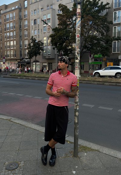 Camisa polo a rayas rojas y blancas, pantalones cortos negros y zapatos negros. Apoyado contra un poste de calle con graffiti, edificios de la ciudad al fondo.