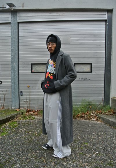 Abrigo largo gris sobre sudadera negra con un gráfico vívido, pantalones anchos grises con rayas blancas y zapatos negros. Entorno urbano con fondo neutro.