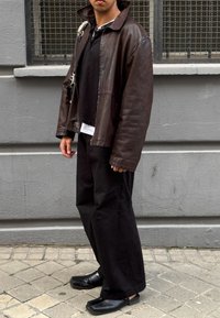 Καφέ δερμάτινο μπουφάν με μεγάλο γιακά, μαύρο πόλο μπλουζάκι, λευκή φανέλα, μαύρα φαρδιά παντελόνια και μαύρα παπούτσια με τετράγωνη μύτη.