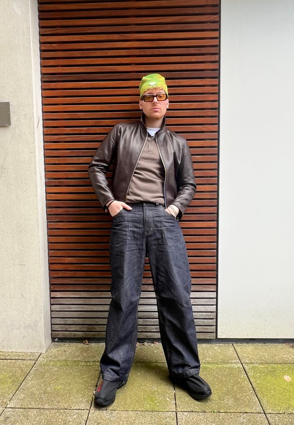 Chaqueta bomber de piel marrón, camisa polo beige, vaqueros oscuros de gran tamaño y zapatos negros. Bandana verde brillante, gafas cuadradas, fondo de madera texturizada.