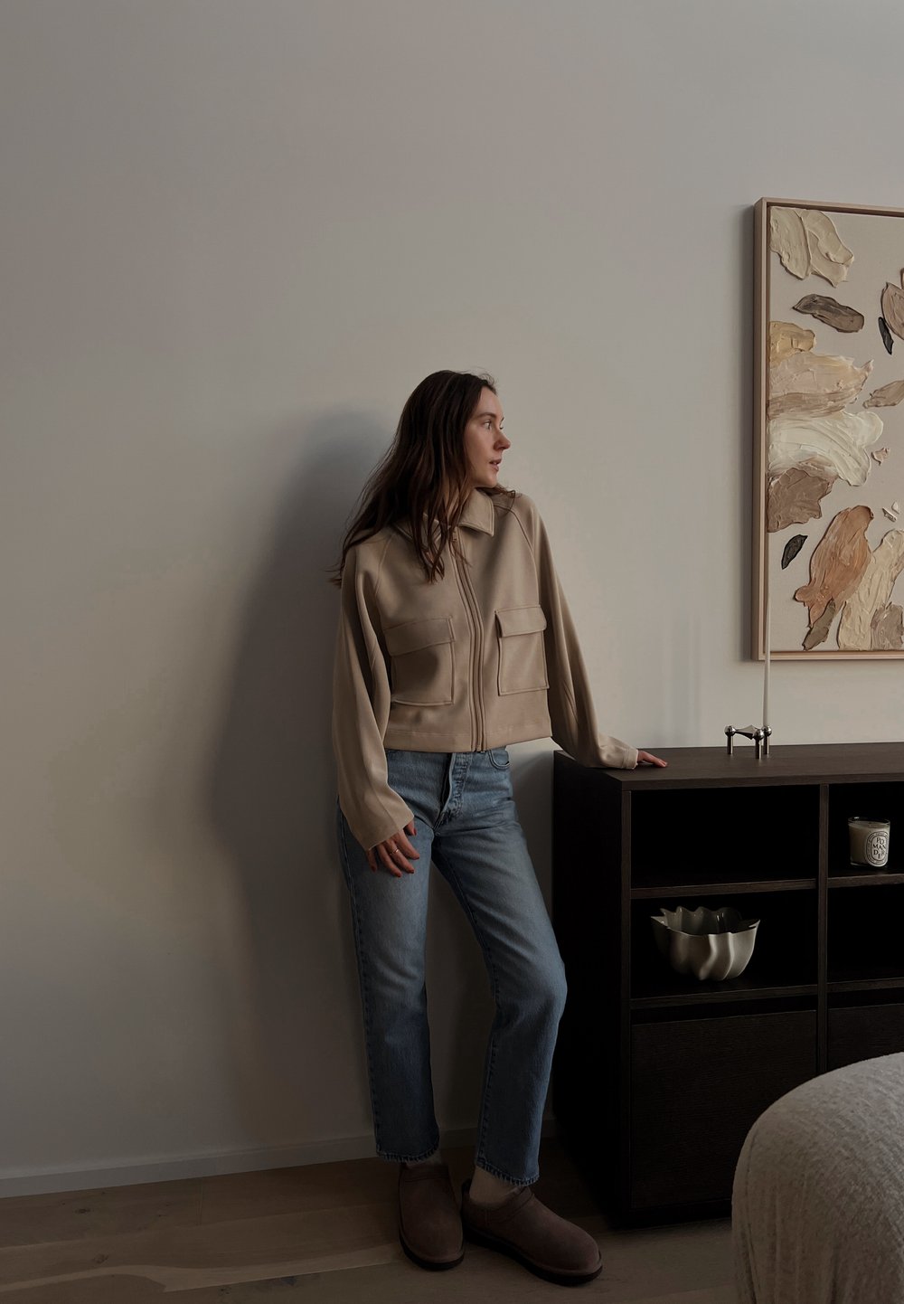 Beež värvi tõmblukuga jakk kahe esitaskuga, helesinised teksad ja pruunid sussid, toetudes tumedast puidust kapile.