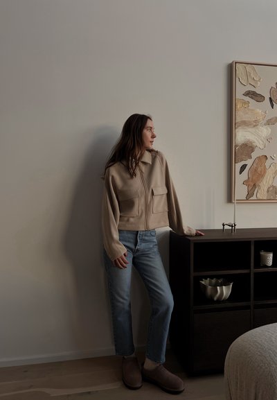 Chaqueta beige con cremallera y dos bolsillos delanteros, vaqueros azul claro y zapatillas marrones, apoyado contra un armario de madera oscura.