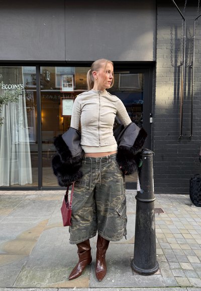 Mujer con top beige con cremallera, pantalones cortos anchos de camuflaje, botas marrones, sosteniendo un abrigo de piel negro y un bolso rojo, de pie en la acera de la ciudad cerca de un poste negro.