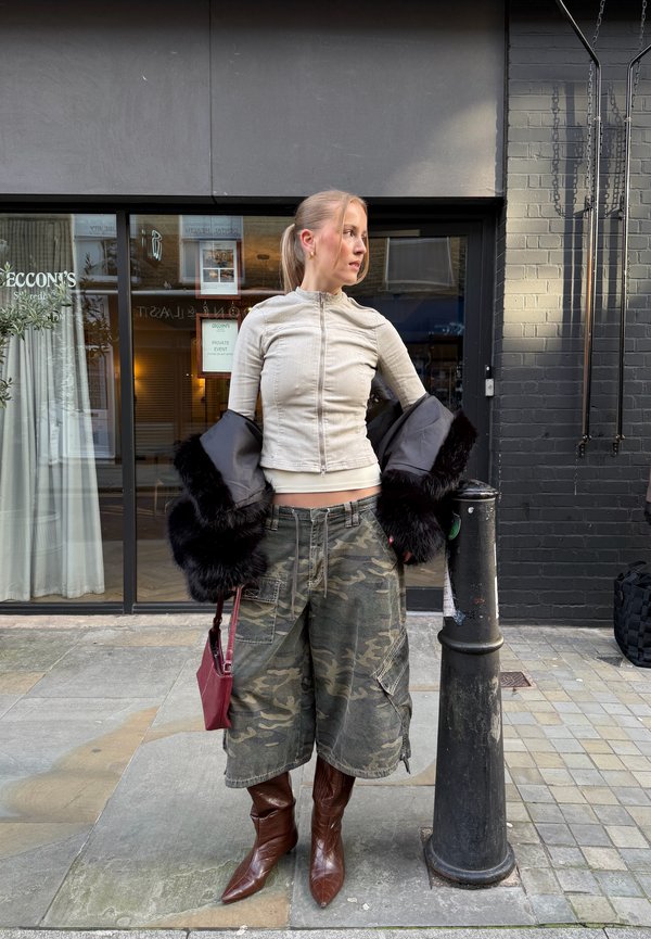 Femme portant un haut beige zippé, un short large camouflage, des bottes brunes, tenant un manteau en fourrure noire et un sac à main rouge, debout sur le trottoir de la ville près d'un poteau noir.