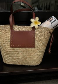 Geweven stro tote bag met bruine leren accenten en gouden studs, met een bloem en een gedeeltelijk zichtbare opgerolde tijdschrift binnenin.