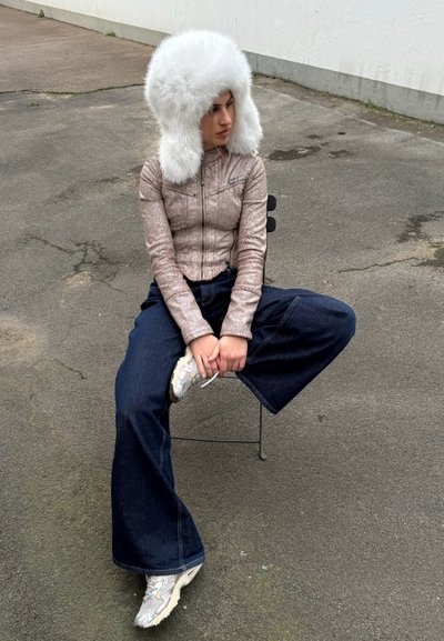 Una modelo que lleva una chaqueta metálica beige con cremallera, unos vaqueros anchos de color azul oscuro y un sombrero blanco esponjoso, sentada en una silla minimalista.