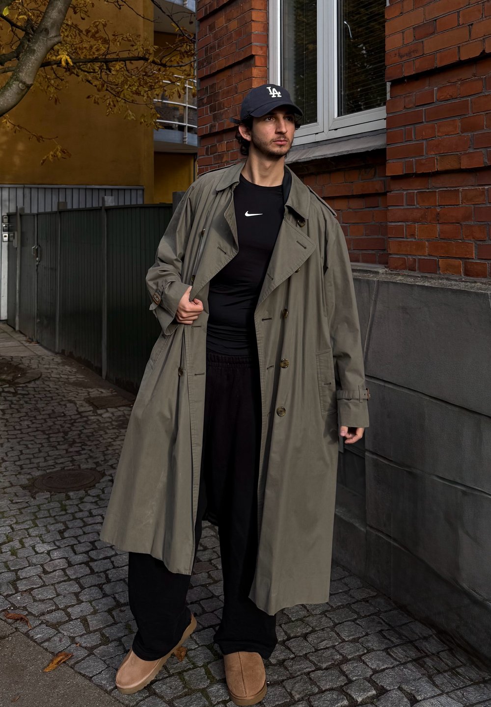 Lang, oliven grønn trenchcoat med avslappet passform, svart treningsskjorte, løse svarte bukser og brune slip-on sko, stående på brostein.