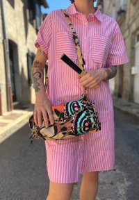 Camisa de botões às riscas em rosa, com dois bolsos no peito; segurando uma mala de mão com padrão multicolorido, adornada com detalhes de atacadores e uma alça destacável.