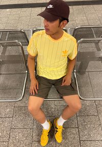 Sportovní outfit skládající se z žlutého pruhovaného trička Adidas, tmavých šortek a žlutých bot Asics s černými akcenty, doplněný o bílé ponožky.