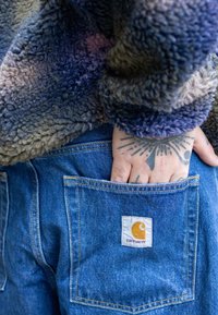 Chlpatá, viacfarebná flísová bunda v odtieňoch modrej a fialovej. Modré džínsové nohavice so zadným vreckom, na ktorom je našitá nášivka s logom Carhartt.