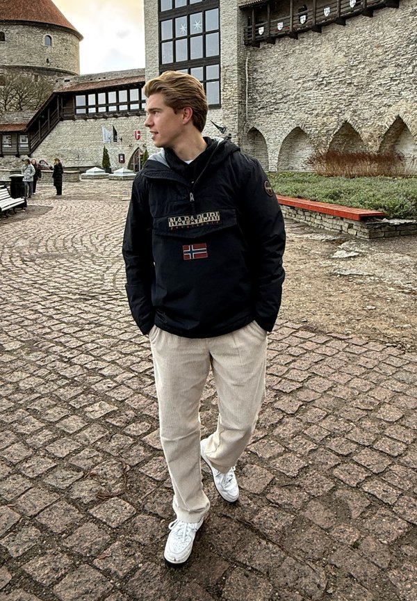 Sort pullover jakke med en forlommet og norsk flagg patch, parret med lyse beige corduroy bukser og hvide sneakers. Stensti og bygninger i baggrunden.