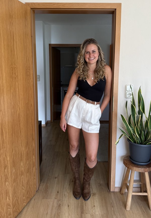 Ung kvinde med krøllet blondt hår smilende, iført en sort top, hvide shorts og brune støvler, stående i en trædøråbning ved siden af en potteplante.