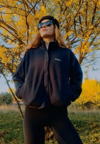 Tamsiai mėlynas flisinis švarkelis su sagų užsegimu ir dviem kišenėmis, dėvimas ant juodų tamprių. Fone matyti geltoni rudeniniai lapai.