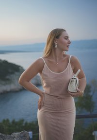Lys rosa satengkjole med spagetti-stropper, kroppsnær silhuett; teksturert kremfarget håndveske holdt i høyre hånd. Scenic kystbakgrunn.