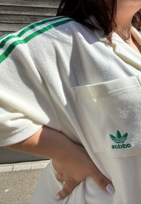 Cilvēks baltā Adidas krekliņā ar zaļām svītrām un logo, roku uz gurniem, brīvā dabā, ar koka sienas fona saules gaismā.