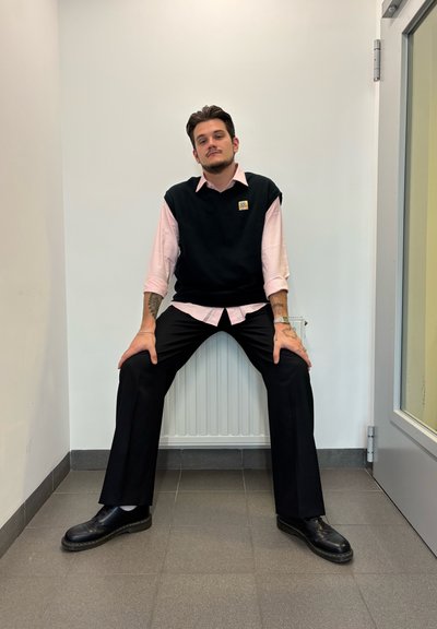 Un suéter negro sin mangas sobre una camisa rosa claro, combinado con pantalones y zapatos negros. Sujeto sentado contra una pared blanca.