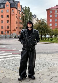 Schwarze Leder-Motorradjacke mit silbernen Reißverschlüssen, kombiniert mit lockeren schwarzen Lederhosen und strukturiertem Schuhwerk, vor einer urbanen Kulisse.