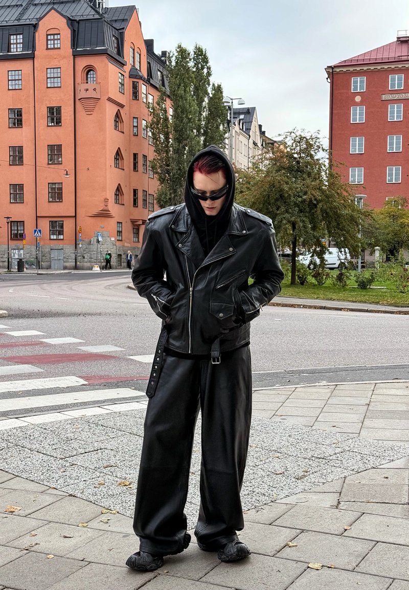 Veste de moto en cuir noir avec des fermetures éclair argentées, associée à un pantalon en cuir noir ample et des chaussures texturées, le tout sur fond urbain.