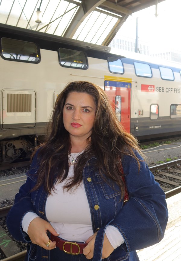 Donna con lunghi capelli castani che indossa una giacca di jeans e una camicia bianca, in piedi sulla banchina accanto a un treno a due piani SBB CFF FFS.