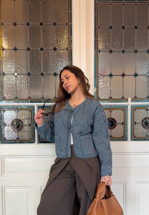 Mujer con chaqueta azul de cuadros y pantalones marrones sostiene una bolsa marrón frente a una puerta de paneles blancos y un vitral.