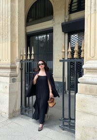 Schwarzes plissiertes Kleid mit strukturiertem Schnitt, kombiniert mit einem schwarzen Umhang und einer braunen Handtasche. Sonnenbrille und ausgefallene Schuhakzente runden den Look ab.