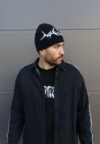 Beanie tricotat negru cu design alb, cămașă din denim cu margini uzate și tricou negru cu text alb. Poartă un cercel.