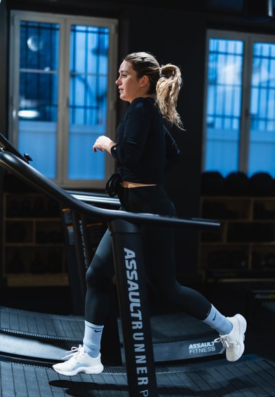 Top negro de manga larga, leggings negros y zapatillas deportivas blancas con calcetines azul claro; corriendo en una cinta de correr Assault Runner negra.