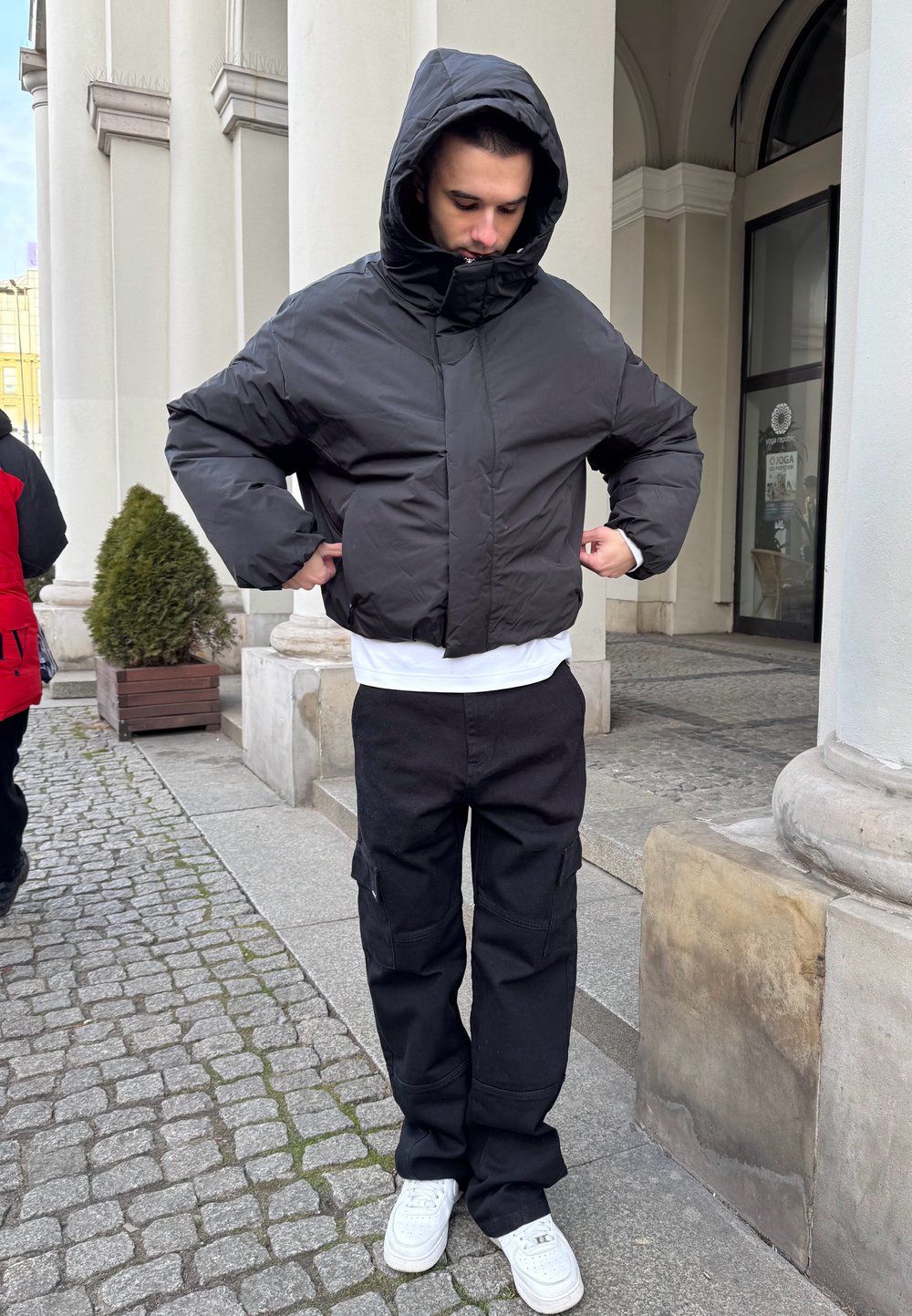 Chaqueta acolchada negra con cuello alto y capucha, combinada con pantalones cargo negros sueltos y zapatillas blancas. Tela texturizada, diseño minimalista.