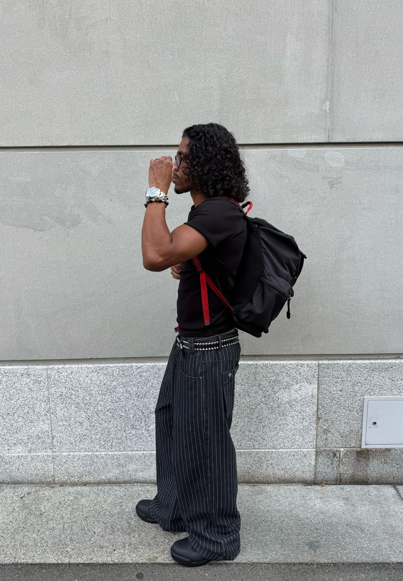 Mochila preta com detalhes em vermelho, camisa preta com camada de textura, calças largas com riscas verticais, e sapatos pretos. Cabelo encaracolado, óculos de sol, e um relógio.