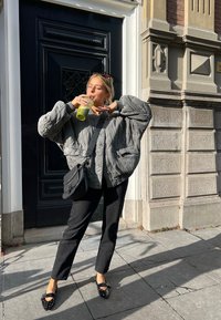 Grau gestepptes Bomberjacke mit oversize Passform, kombiniert mit schwarzen gerade geschnittenen Hosen und mit Nieten verzierten schwarzen Ballerinas. Hält ein grünes Getränk.