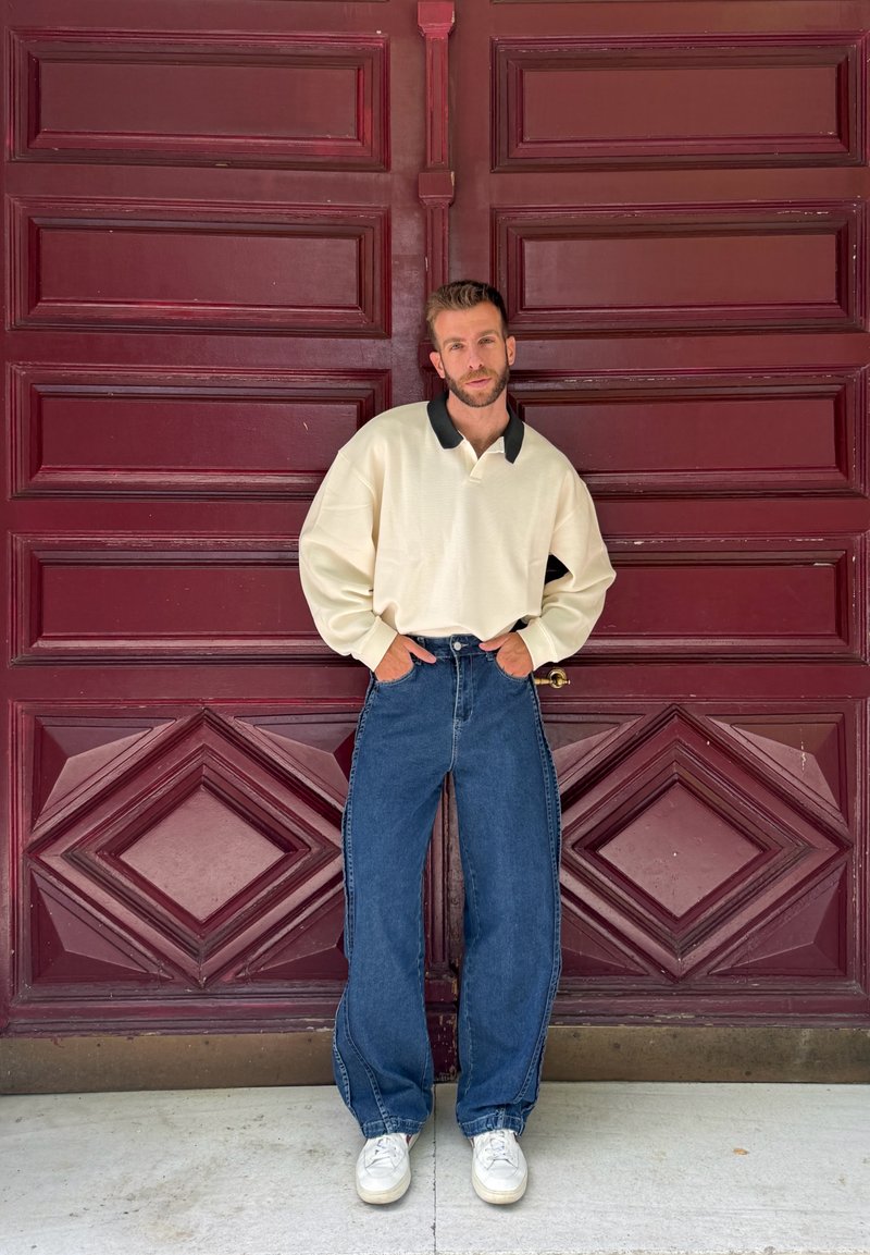 Maglione crema con colletto nero, abbinato a jeans blu larghi e sneaker bianche, posizionato davanti a una porta geometrica color bordeaux.