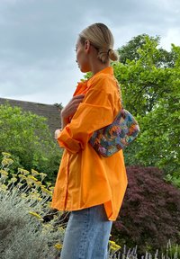 Saco de mão com pérolas e borlas coloridas, apresentando uma alça de corrente. Usado com uma camisa oversized laranja brilhante e jeans de denim.