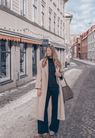 Mujer con abrigo beige y conjunto azul marino, sosteniendo una bolsa marrón, de pie en una calle empedrada con bordes nevados cerca de escaparates.