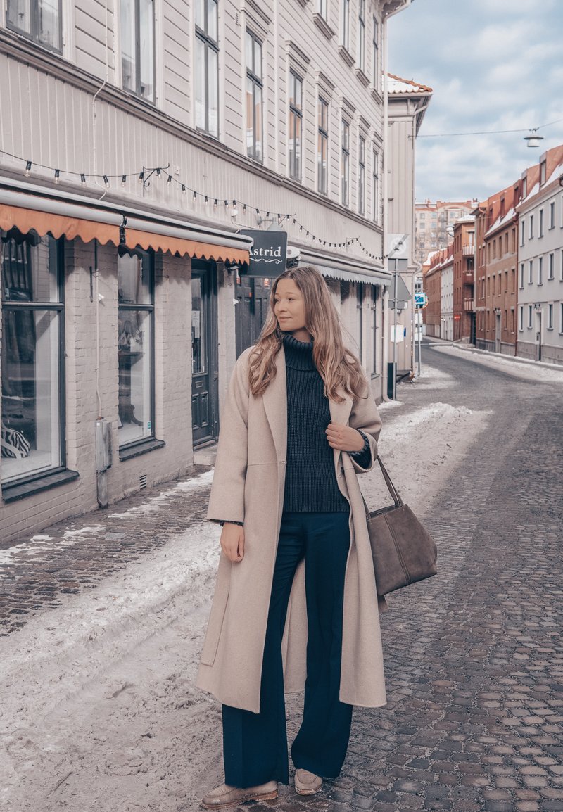 Femme en manteau beige et tenue bleu marine tenant un sac marron, debout dans une rue pavée aux bordures enneigées près des devantures de magasins.