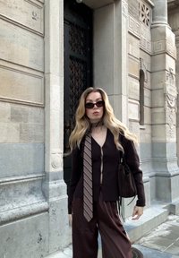 Brūns adīts kardigans ar pogu aizdari, kombinēts ar saskaņotām vaļīgām biksēm, svītrainu kaklasaiti un mazu tumšu somiņu. Tumšas saulesbrilles noslēdz izskatu.