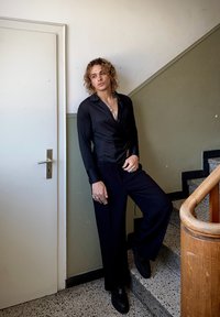 Sort satin langærmet skjorte med V-udskæring, parret med brede sorte bukser. Dekorative ringe og lagdelte halskæder. Lys baggrund.
