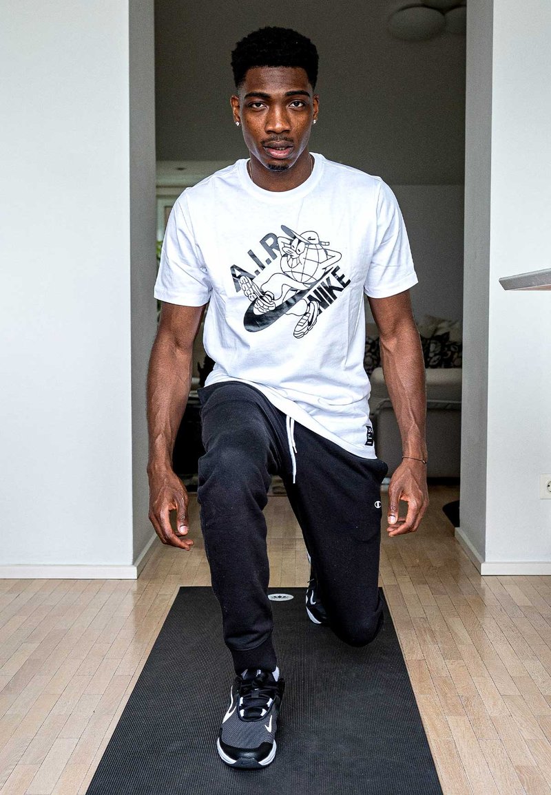 White t-shirt with black graphic and text, black sweatpants, and black athletic shoes. The individual is kneeling on a yoga mat.