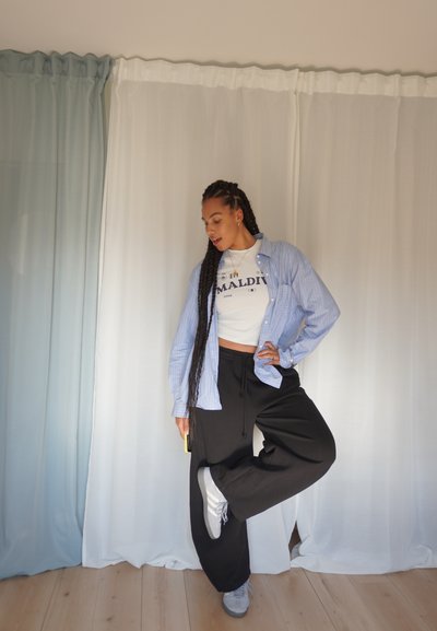 Pantalones deportivos negros holgados, camiseta corta gráfica blanca y camisa de botón a rayas azul claro. Zapatillas gris sobre un suelo de madera clara. Fondo liso.