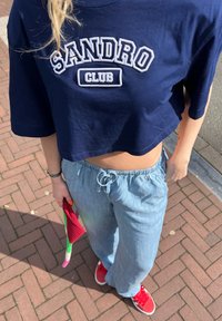 Marinblå croppat topp med "SANDRO CLUB" text, ljusa denimbyxor med elastisk midja, röda sneakers, håller en färgglad virkade väska.