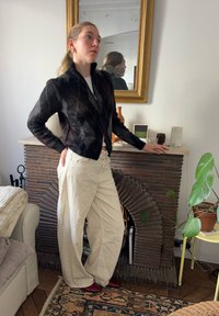 Črna jakna z zadrgo iz teksturirane tkanine, krem hlače širokih hlačnic in rdeči čevlji. Rjavi poudarki v prijetnem notranjem prostoru z rastlinami.