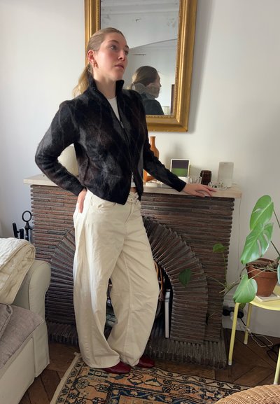 Chaqueta negra con cremallera y tela texturizada, pantalones anchos color crema y zapatos rojos. Detalles marrones en un ambiente acogedor con plantas.