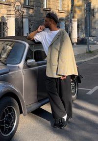Muškarac u svijetlom houndstooth sako preko bijelog majice, crnim širokim hlačama, crnim cipelama i beretkom, naslonjen na sivi vintage automobil.