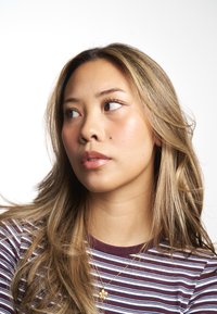 Une femme aux longs cheveux ondulés châtain clair, portant une chemise rayée bordeaux et un collier avec un pendentif en forme de fleur dorée. Fond blanc uni.